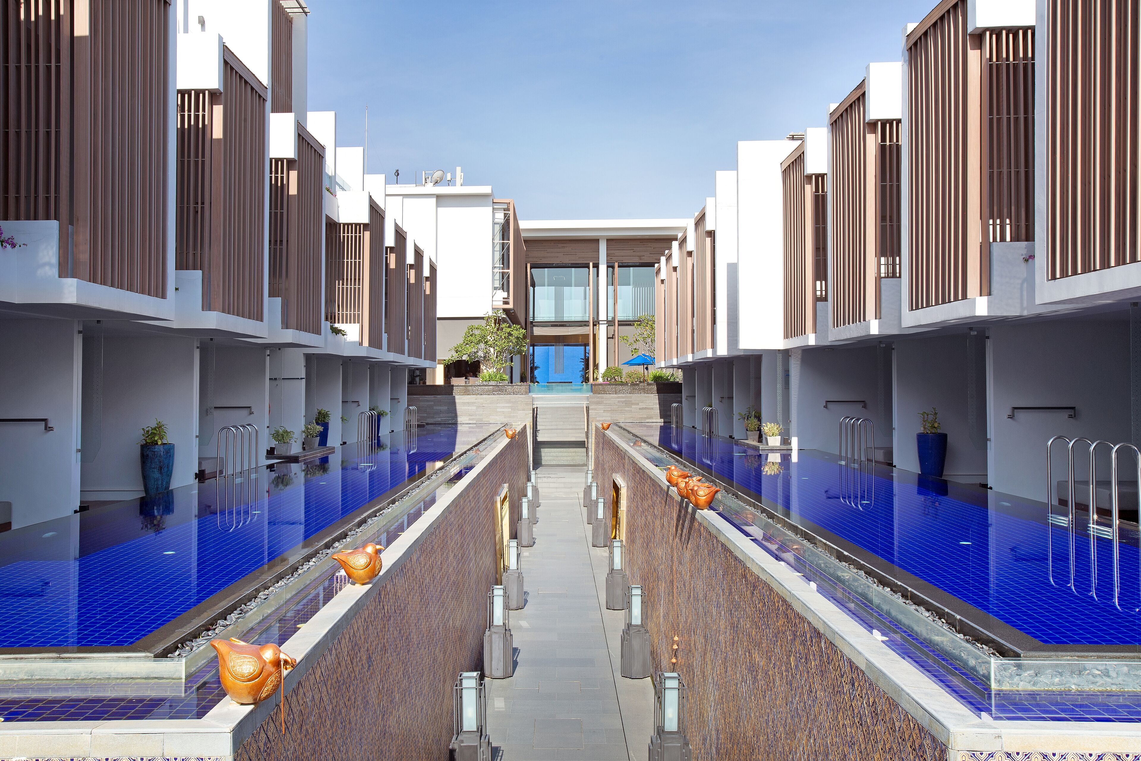 Un hotel moderno con dos piscinas, un paseo central y un edificio con listones verticales de madera.