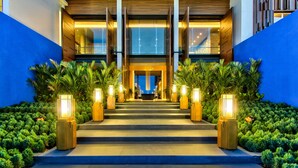 A hotel entrance with a staircase, illuminated by tall, rectangular lights, flanked by lush greenery and blue walls.
