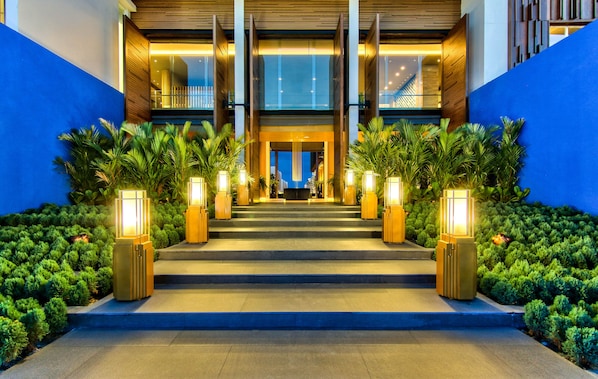 L’entrée d’un hôtel, avec un escalier illuminé par de grands luminaires rectangulaires, bordée d’une végétation luxuriante et de murs bleus.