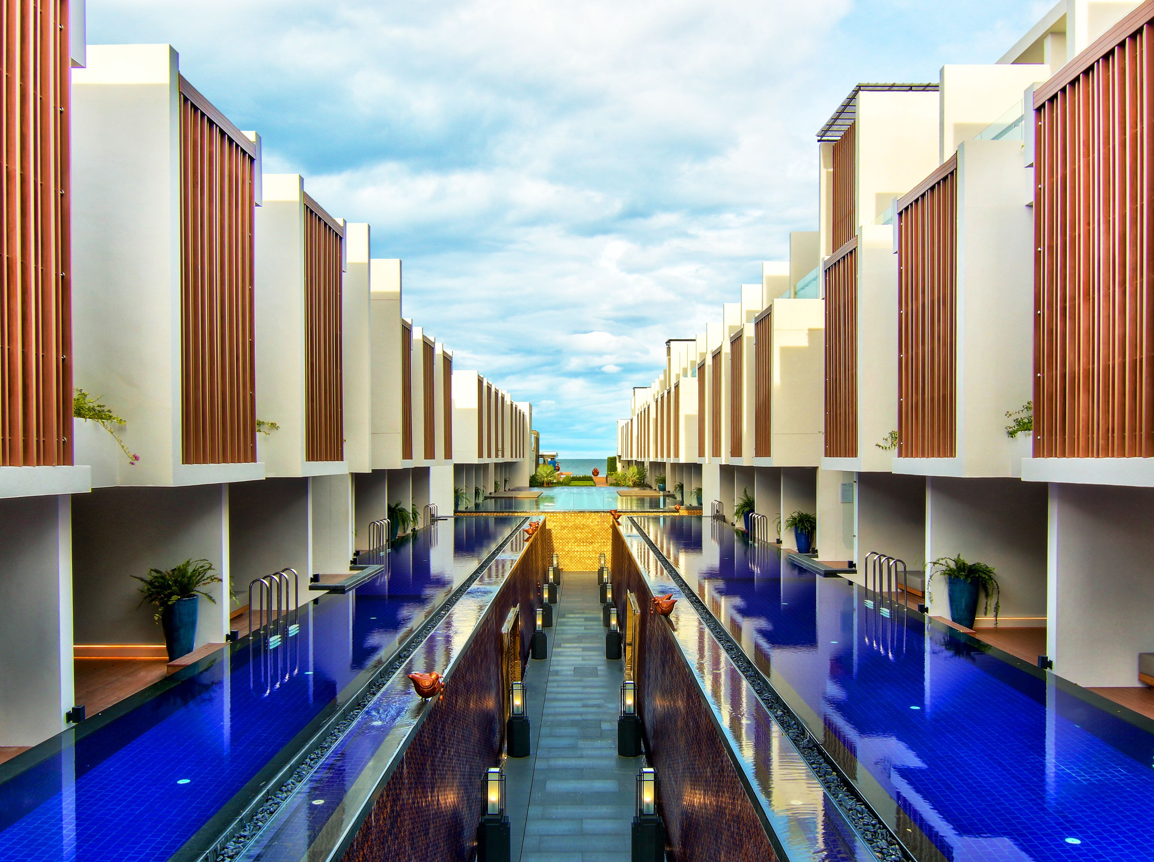 Un hôtel moderne doté d’une piscine centrale, flanqué de deux ailes, avec un ciel dégagé et une vue sur la mer.