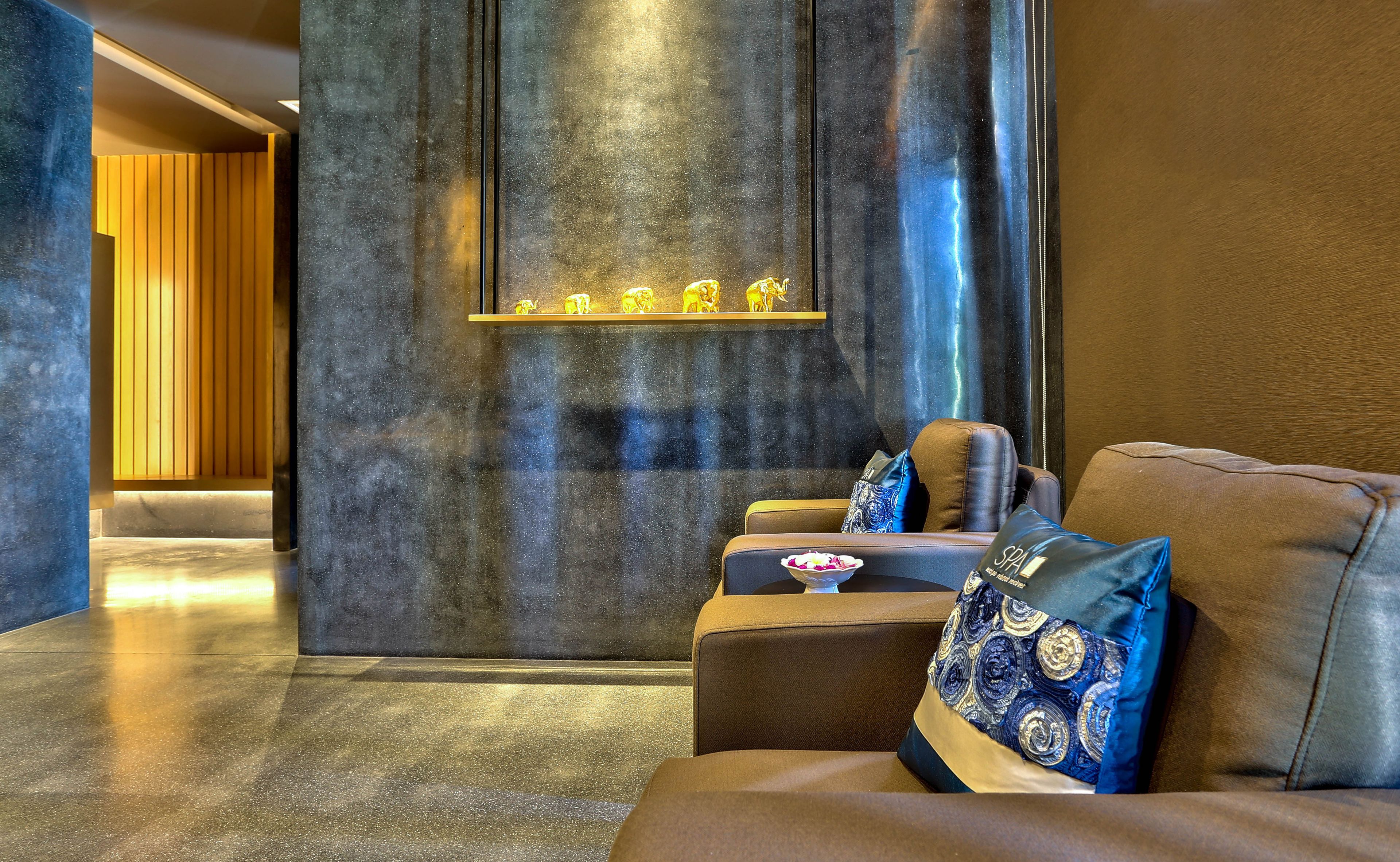 A modern spa area with a dark textured wall, a shelf with golden animal figurines, and a brown leather sofa with patterned cushions.