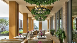 A modern outdoor dining area with wooden furniture, a central chandelier, and large windows offering views of the surrounding landscape.