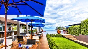 A seaside restaurant with outdoor seating, blue umbrellas, and a view of the ocean.
