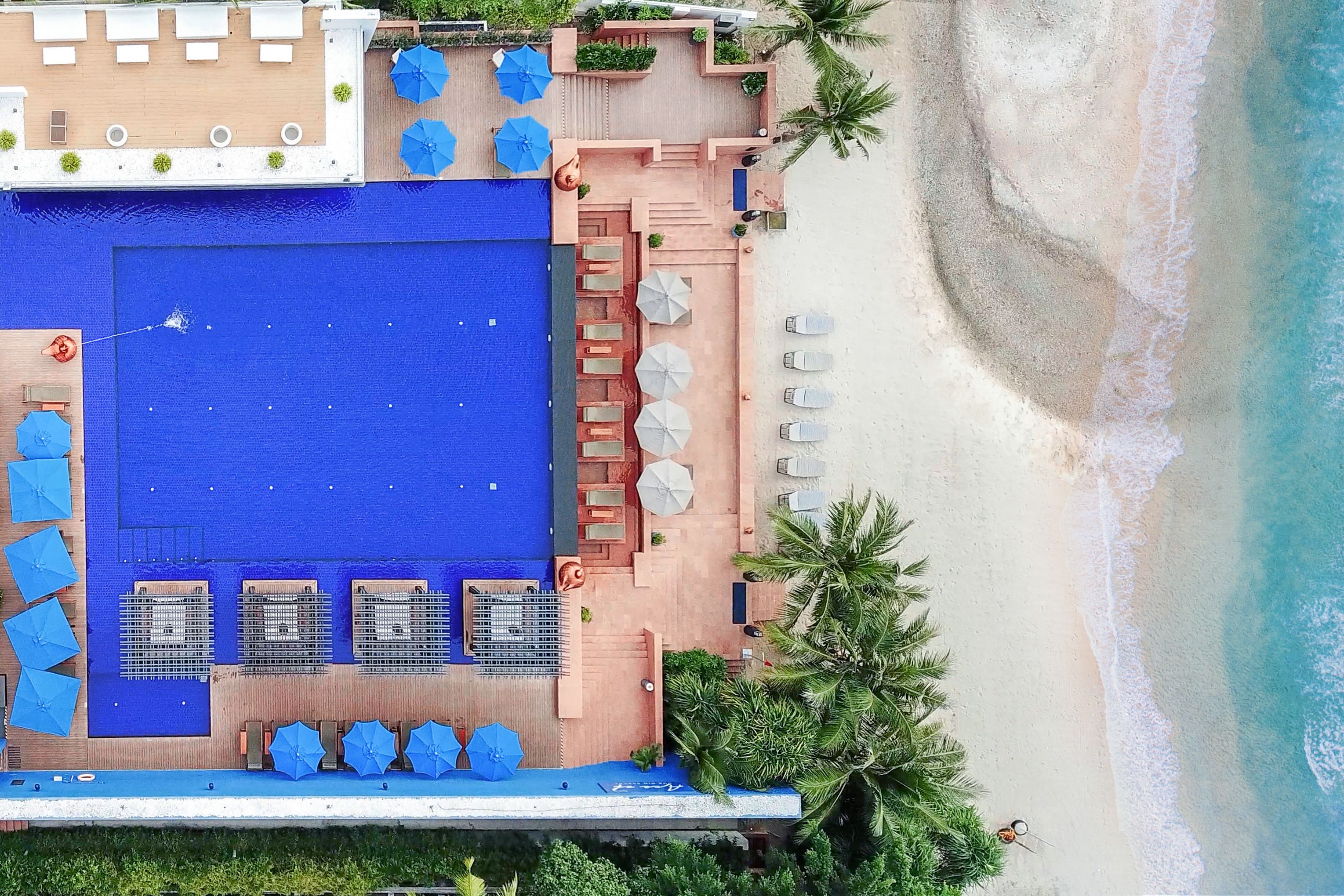An aerial view of a resort with a large blue pool, sun loungers, and a beach area.