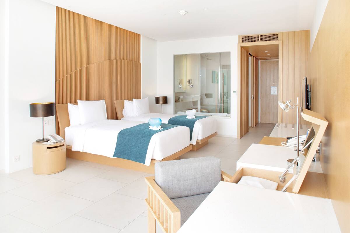 Ein modernes Hotelzimmer mit zwei Betten, einem Schreibtisch, einem Sessel, einem Spiegel und einem Fernseher.