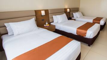 Habitación de hotel con tres camas, cada una con ropa blanca y cabeceros marrones. Hay mesitas de noche con lámparas y un pequeño objeto decorativo en una de ellas.