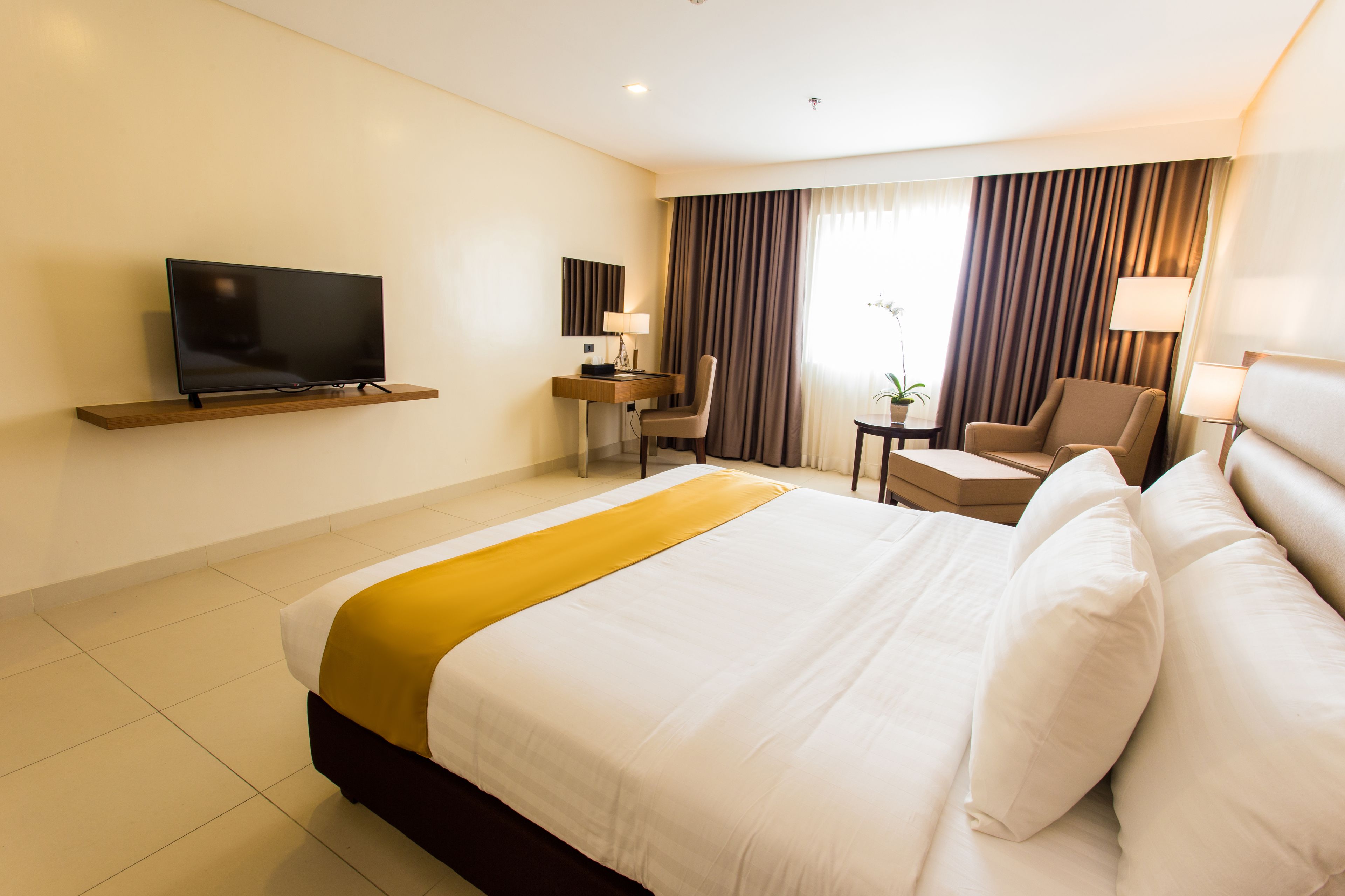 Habitación de hotel con una cama grande, un televisor montado en la pared, un escritorio con una silla y una ventana con cortinas.