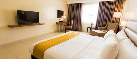 Habitación de hotel con una cama grande, un televisor montado en la pared, un escritorio con una silla y una ventana con cortinas.