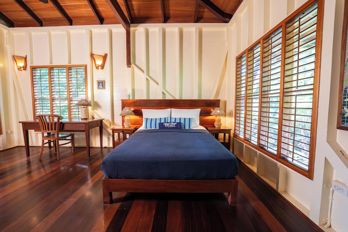 A bedroom with a wooden bed, a desk, and two chairs. There are windows with blinds and wall-mounted lamps.