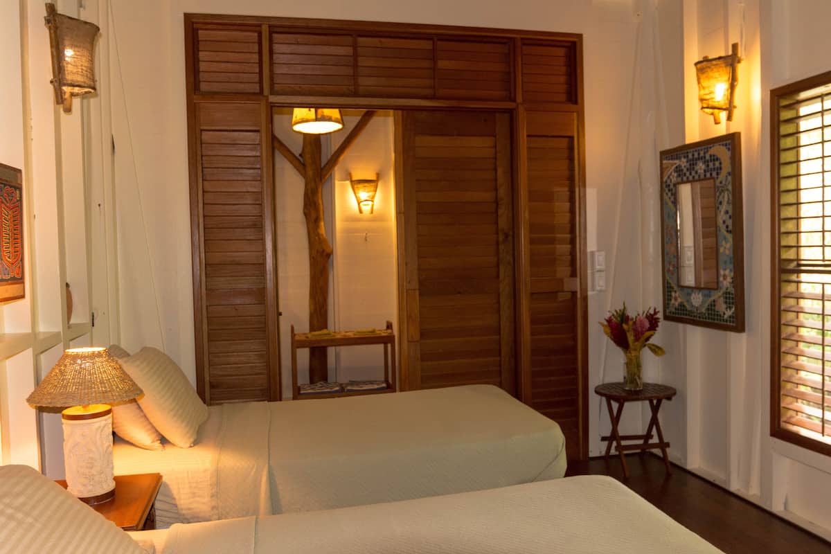 A hotel room with two beds, a wooden wardrobe, a small table with a lamp, and a mirror on the wall.