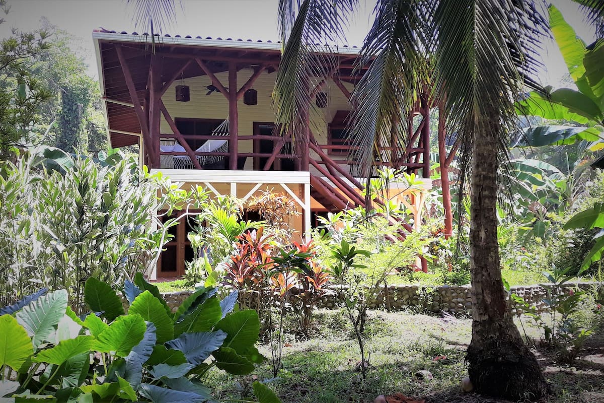 A building with a wooden structure and a covered entrance, surrounded by lush greenery and plants.