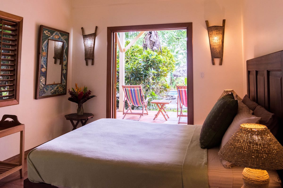 A bedroom with a bed, a nightstand, a mirror, a window, and a view of the outside.