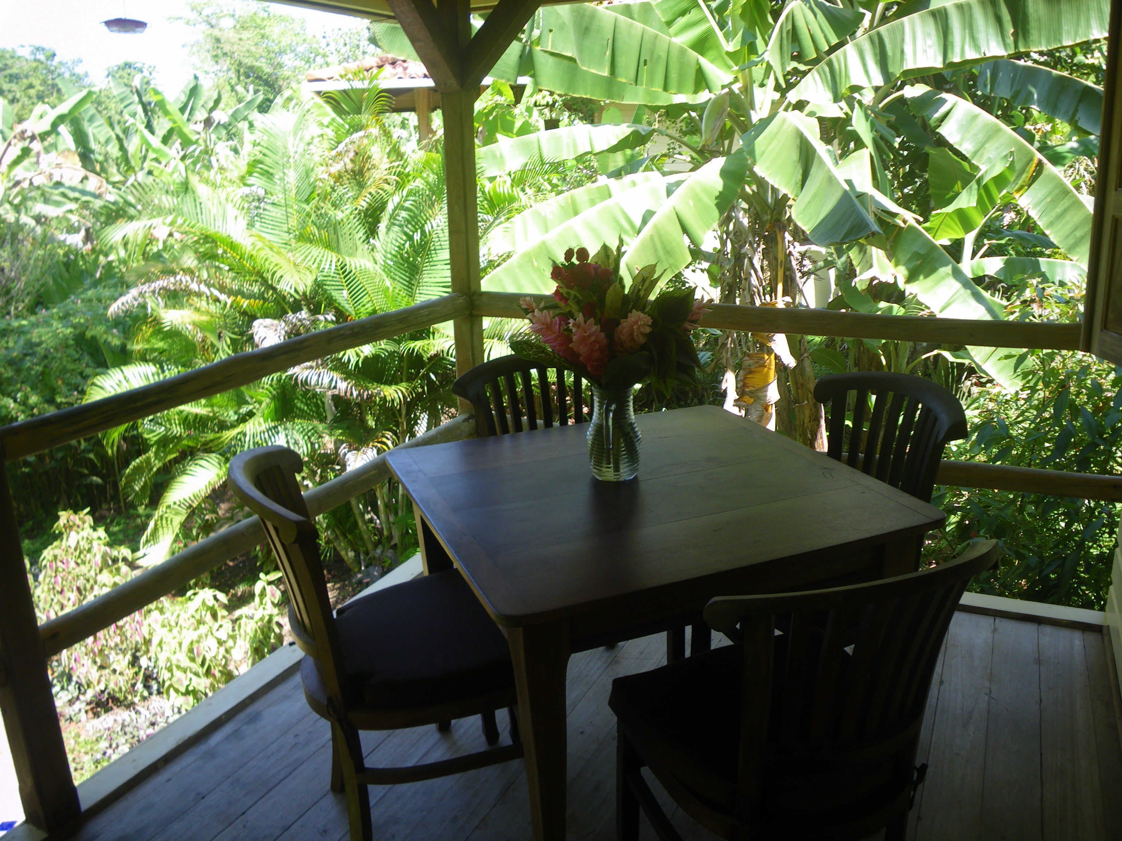 Une table en bois, entourée de chaises, et un vase rempli de fleurs, le tout installé sur un balcon offrant une vue sur une végétation luxuriante.