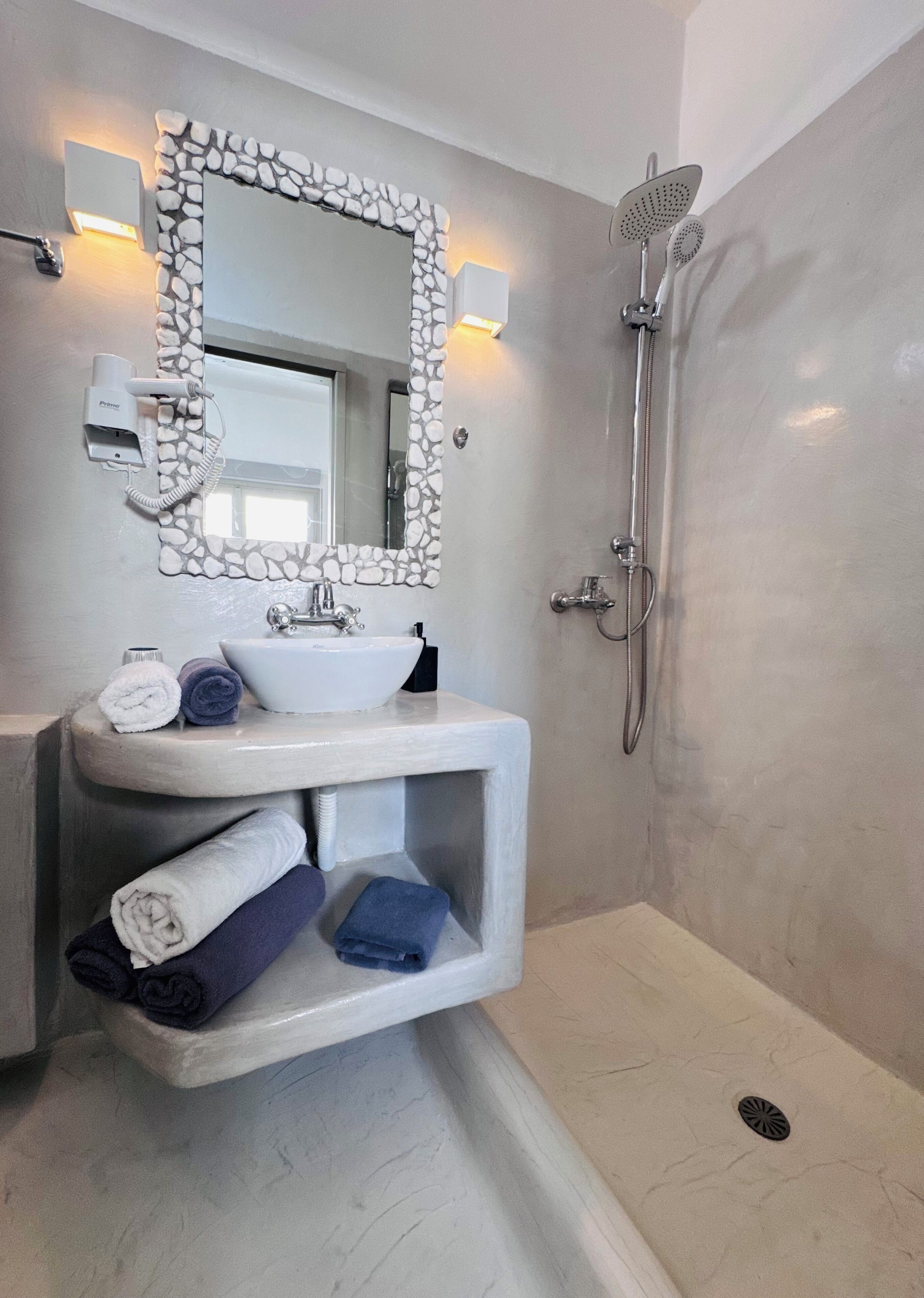 Une salle de bain avec un miroir encadré de pierre, un lavabo avec un receveur blanc et une douche équipée d’un pommeau de douche à main.