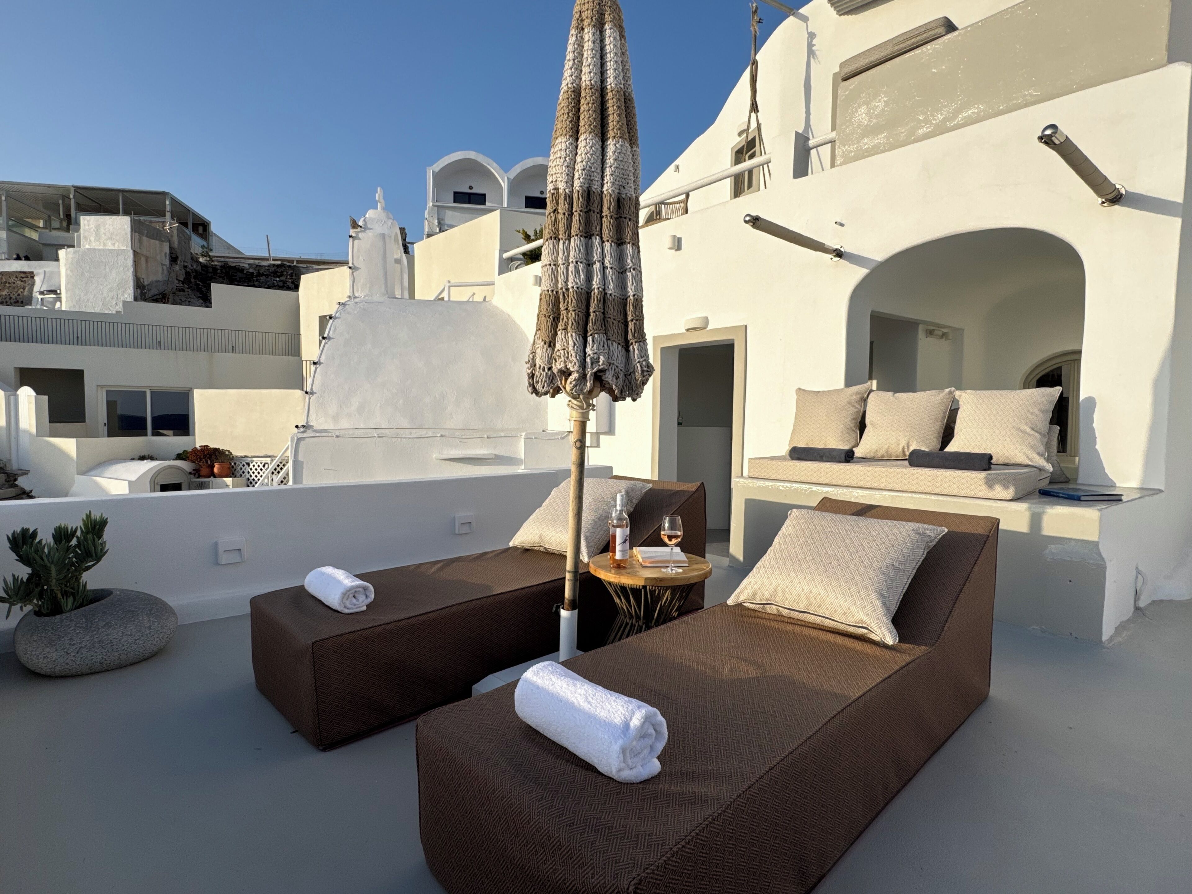 Eine Dachterrasse mit zwei Liegestühlen, einem kleinen Tisch mit Flasche und Glas sowie einem großen Sonnenschirm.