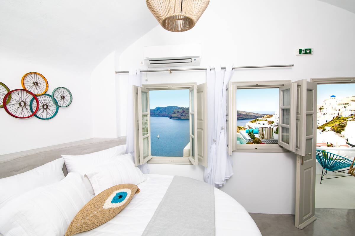 Een slaapkamer met een bed, uitzicht op zee en een raam met uitzicht op een wit gebouw.