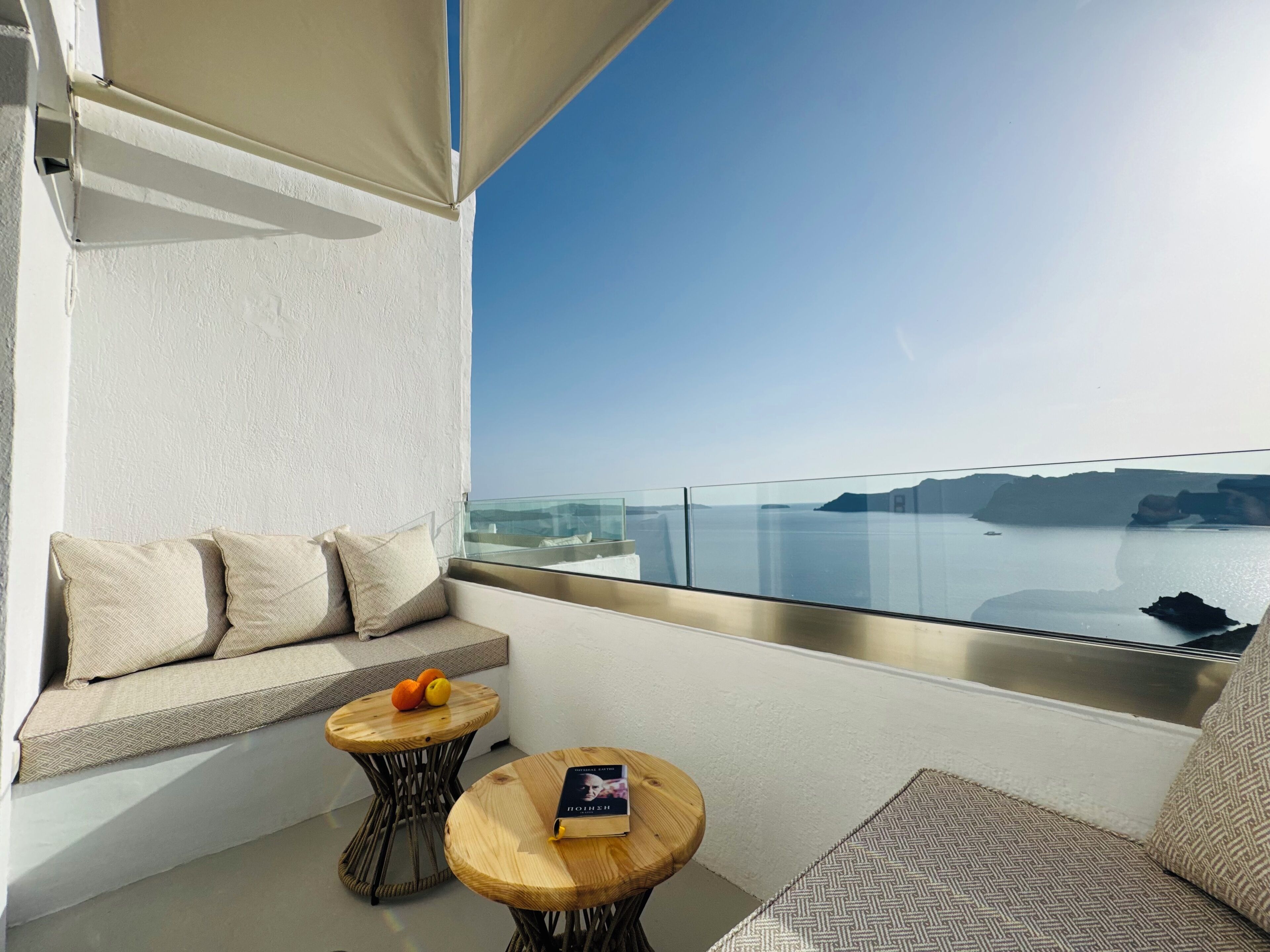 A balcony with a sofa and two round tables, one with a book and oranges, and a clear view of the sea and distant land.