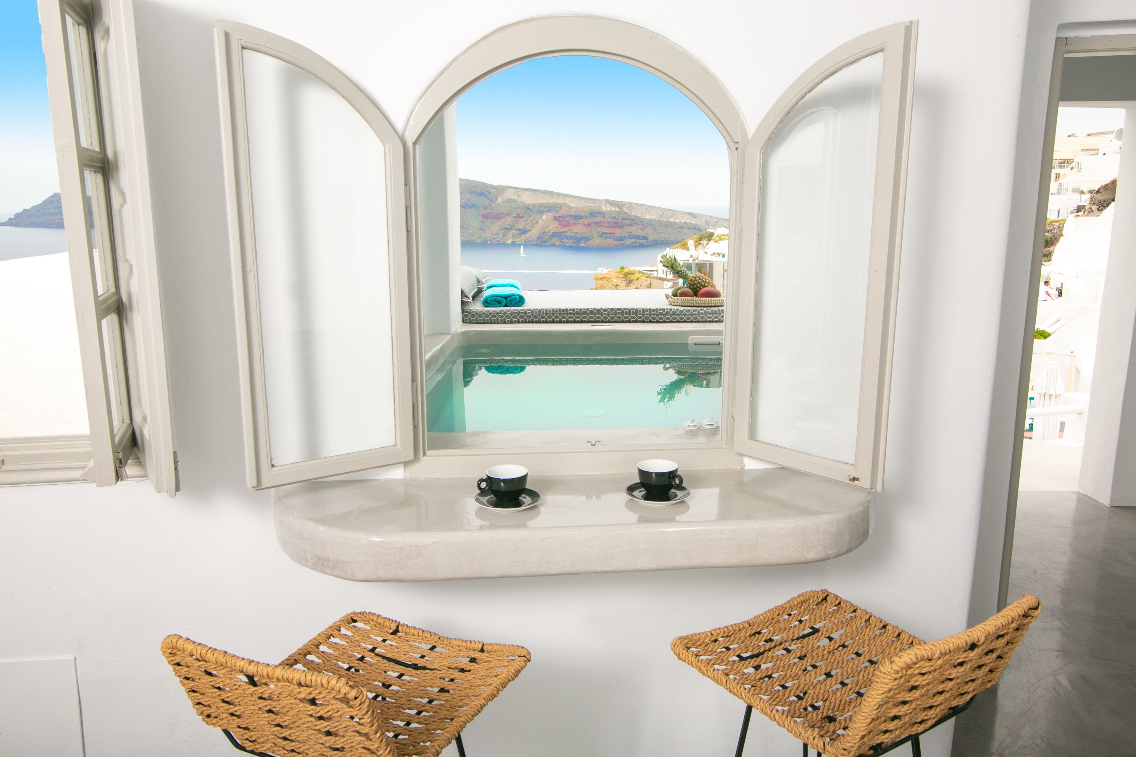 Ein Zimmer mit Blick auf ein Schwimmbad und Berge, zwei Korbstühle und ein kleiner Tisch mit zwei Tassen.