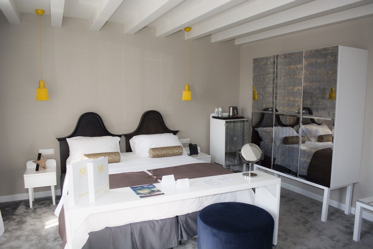 A hotel room with a bed, bedside tables, a desk, a chair, a wardrobe, and a mirror.