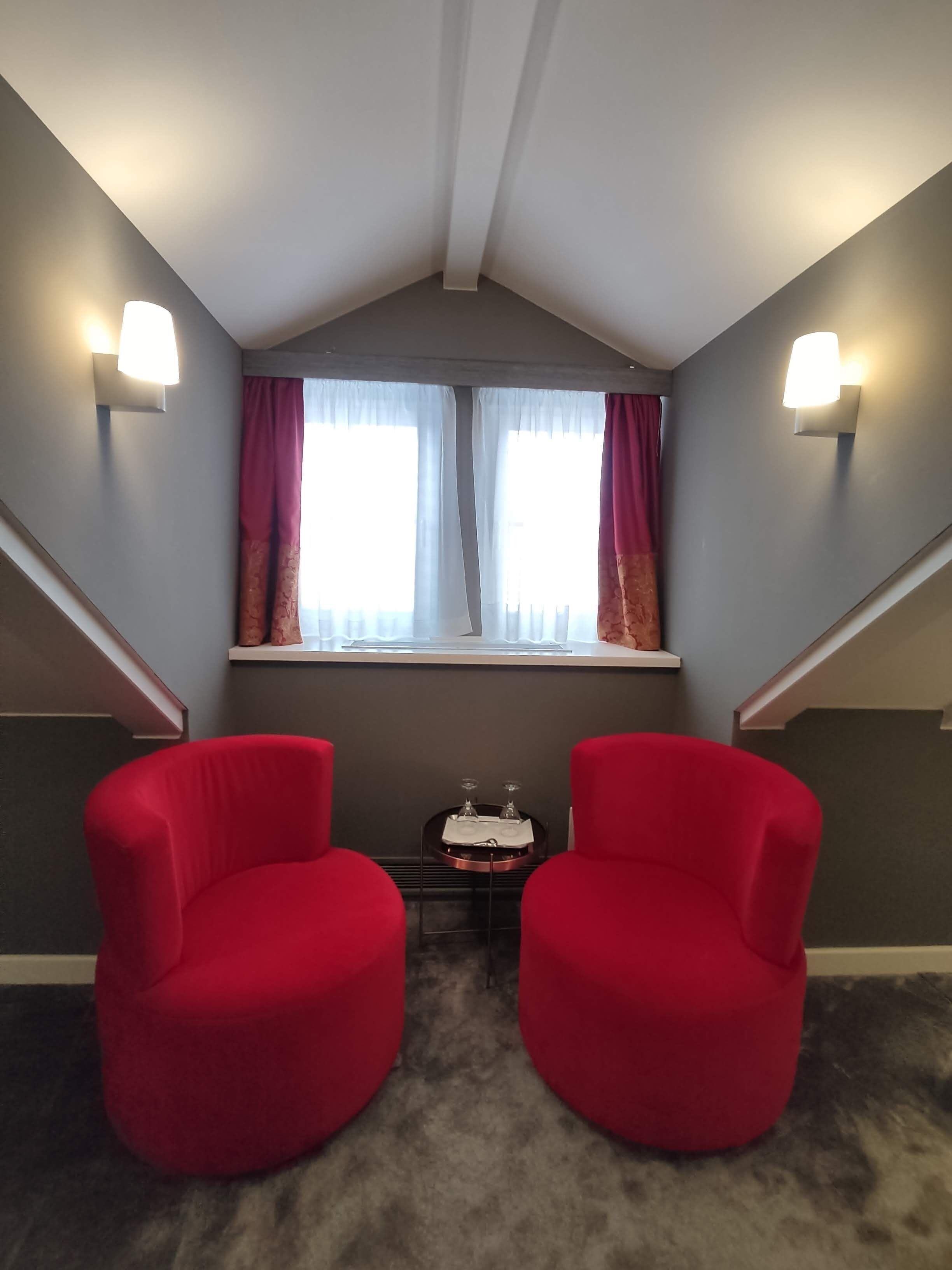 Una habitación con techo inclinado, dos sillas rojas, una mesita con bandeja y una ventana con cortinas.