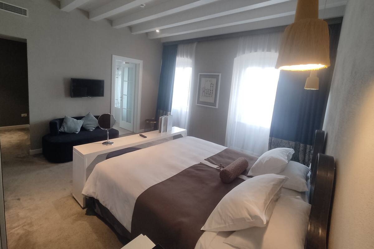 A modern hotel room with a large bed, a bedside table, a TV mounted on the wall, and a ceiling light fixture.