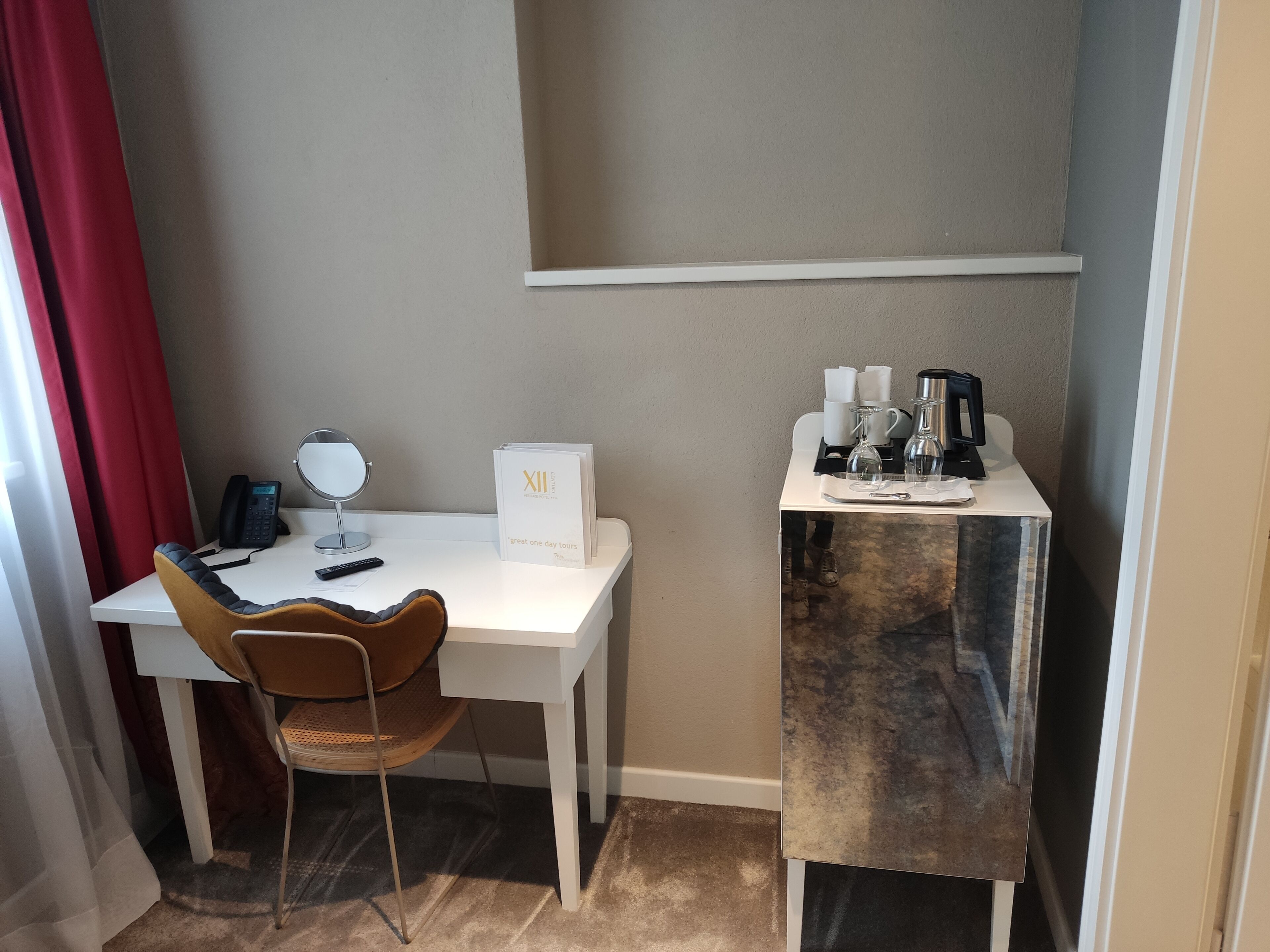 Habitación de hotel con un escritorio blanco, una silla, un espejo, un teléfono, una mesita con café y té, y una pequeña mesa con botellas.