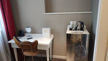 Habitación de hotel con un escritorio blanco, una silla, un espejo, un teléfono, una mesita con café y té, y una pequeña mesa con botellas.