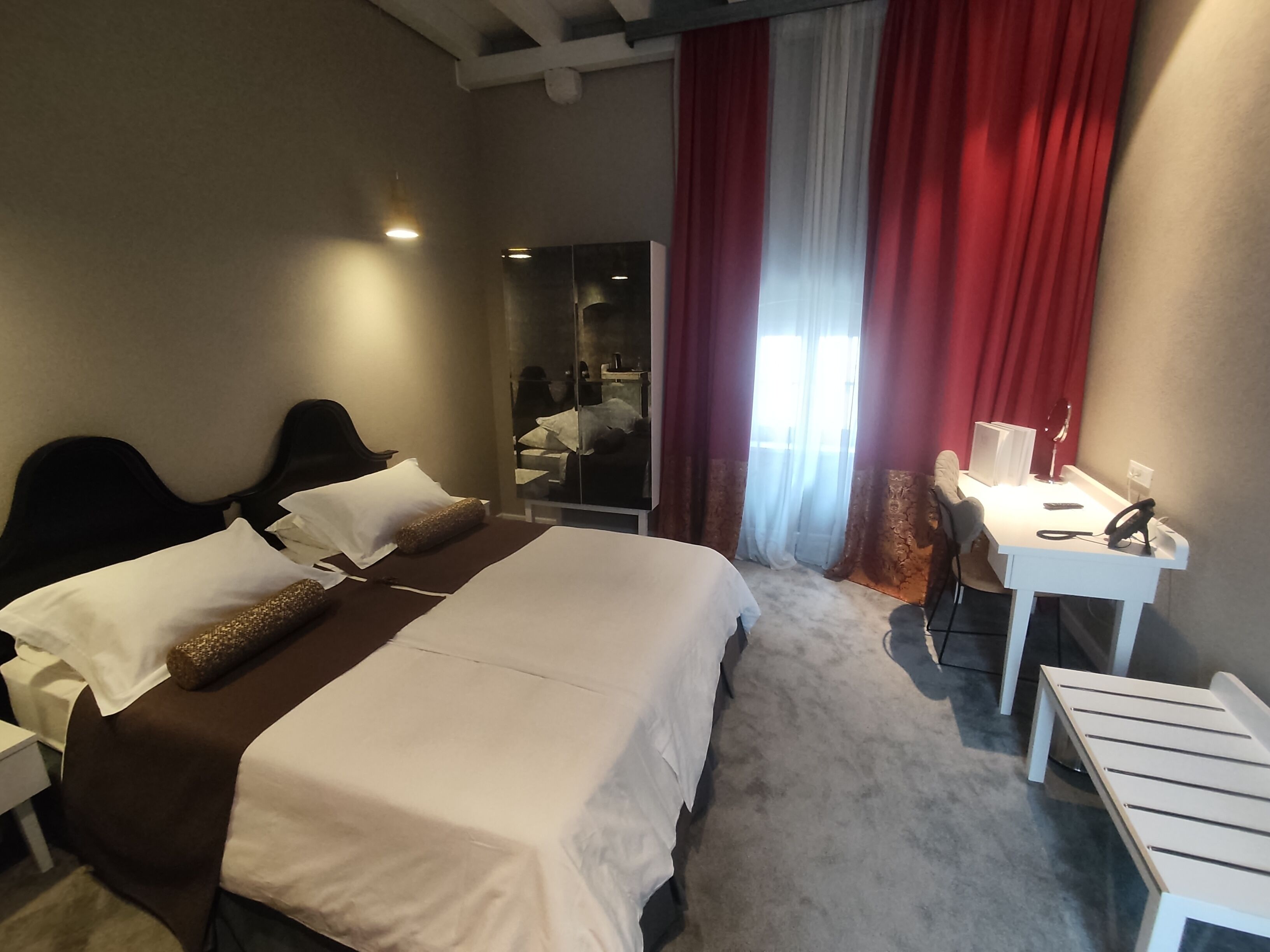 A hotel room with a large bed, a desk, and a chair. There is a mirror and a window with red curtains.
