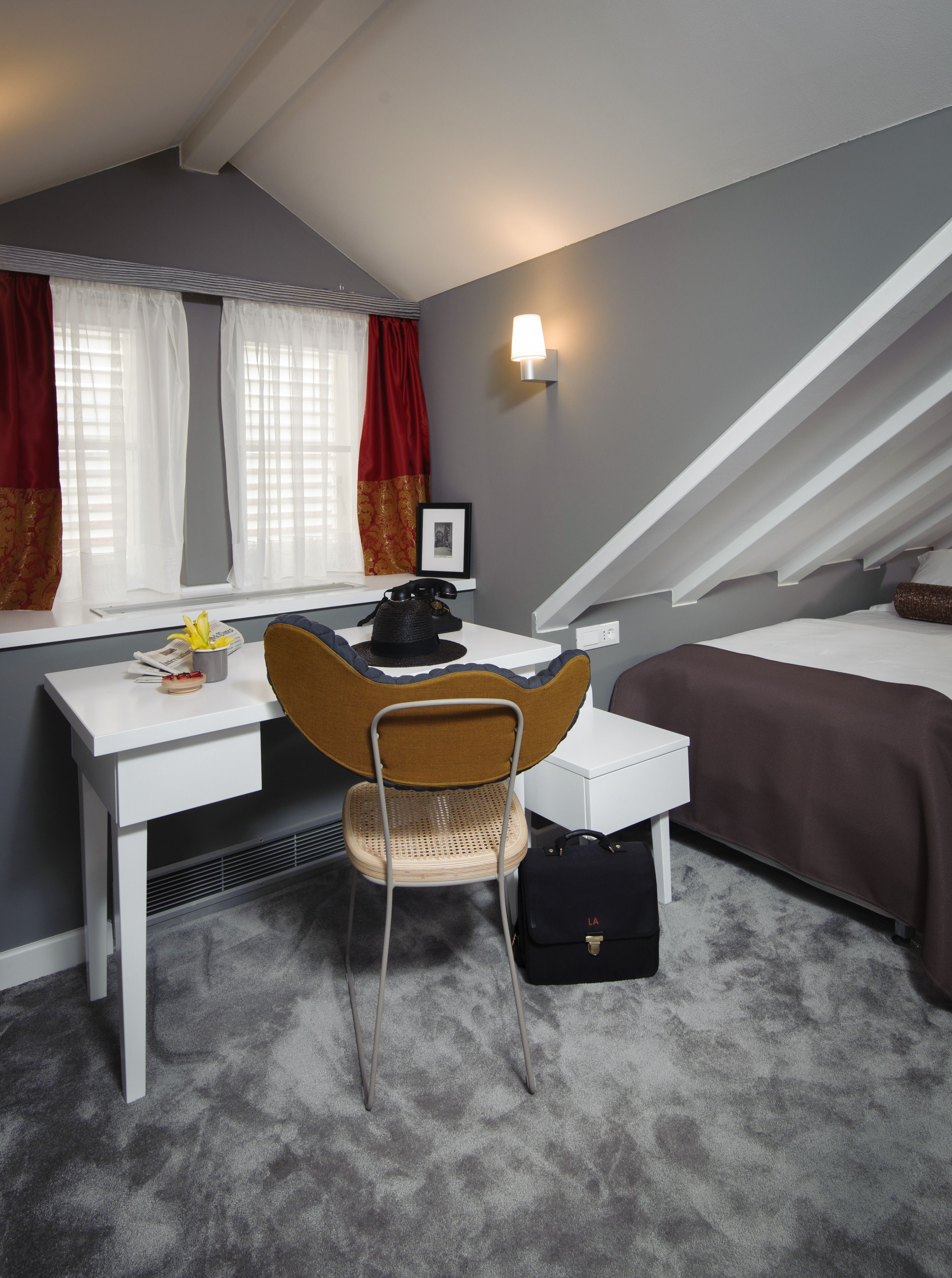 Una habitación de hotel compacta con un escritorio blanco, una silla, una cama con cubrecama marrón y una ventana con cortinas rojas.