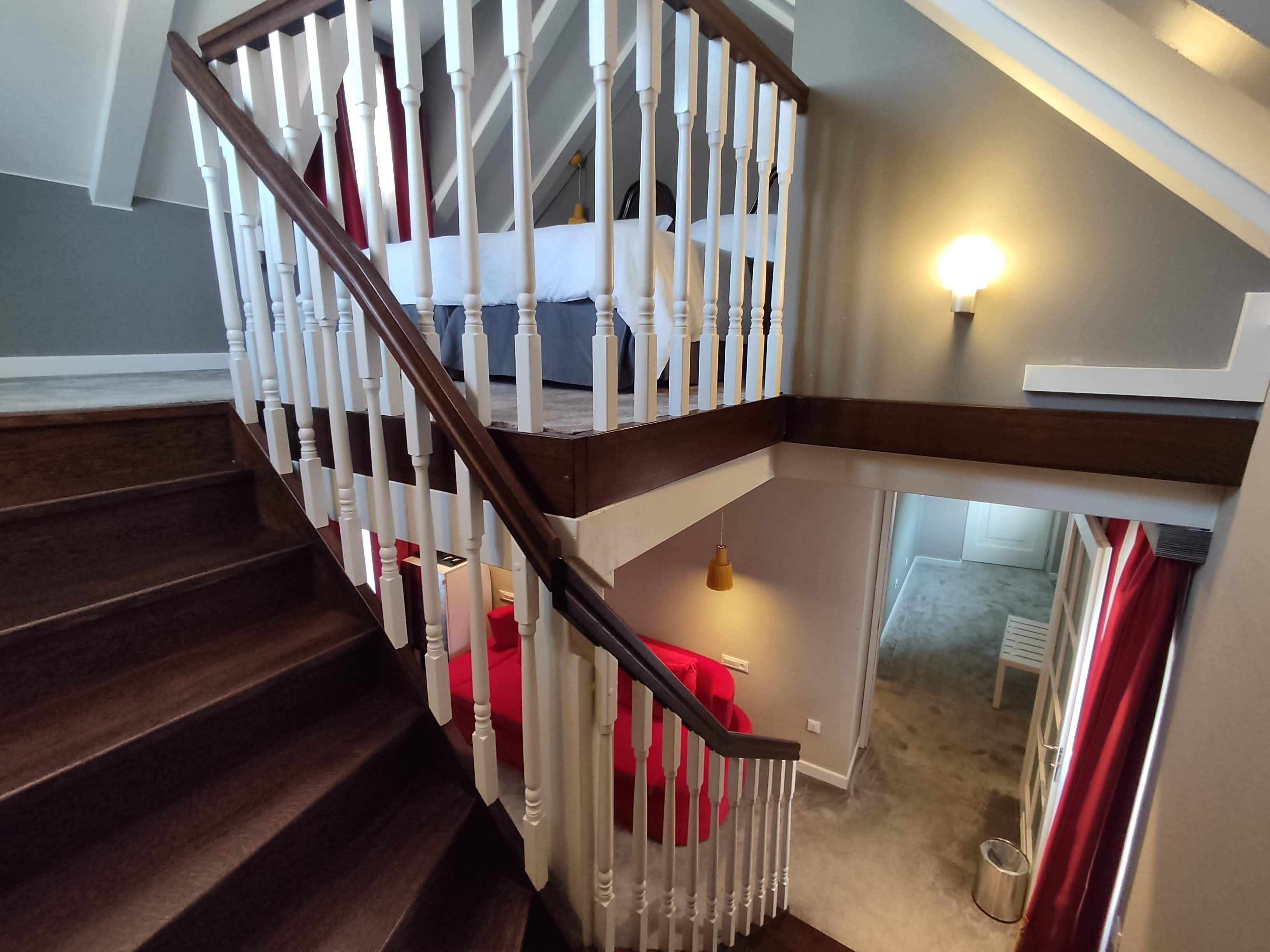 A wooden staircase with white balusters and a red handrail, leading to a landing with a red chair and a hanging pendant light.