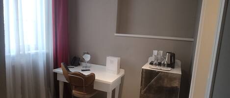 A small, neatly arranged room with a white desk, a chair, a small table with a mirror, a coffee maker, and a bookshelf.