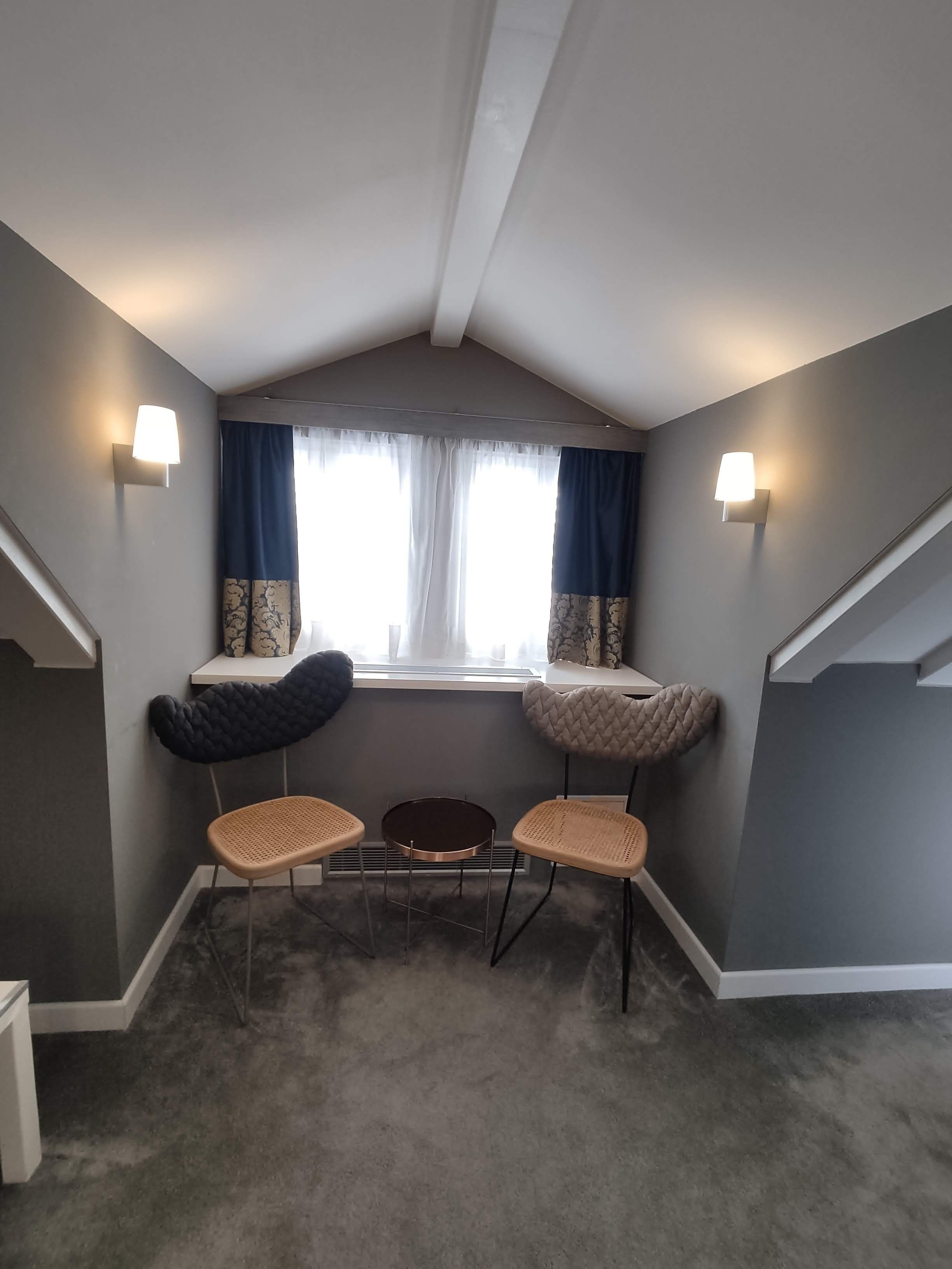 A small, cozy room with a sloped ceiling, two cushioned chairs, a small round table, and a window with curtains.