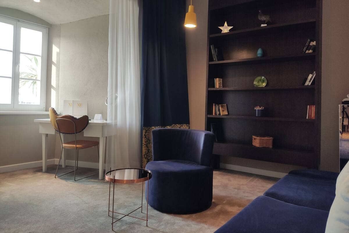 A modern living room with a round coffee table, a blue armchair, a dark bookshelf, and a desk with a chair.