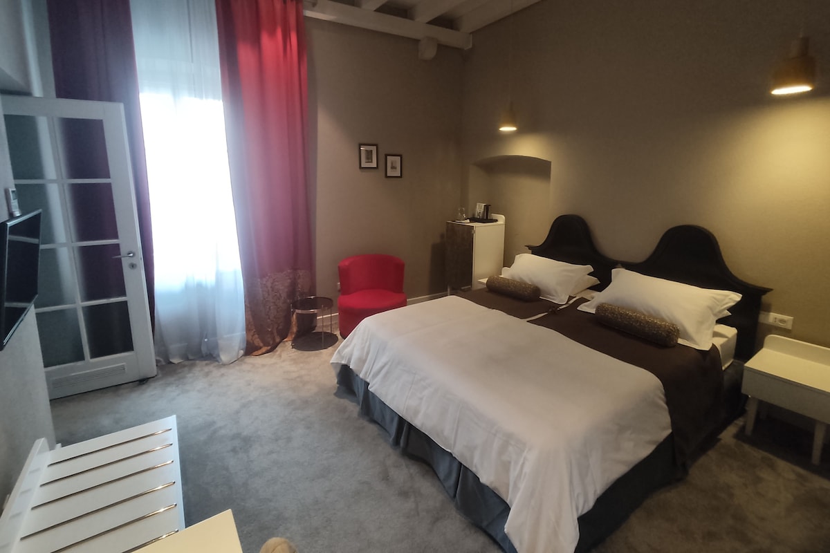 A bedroom with a bed, a red chair, a bedside table, and a door leading to another room.