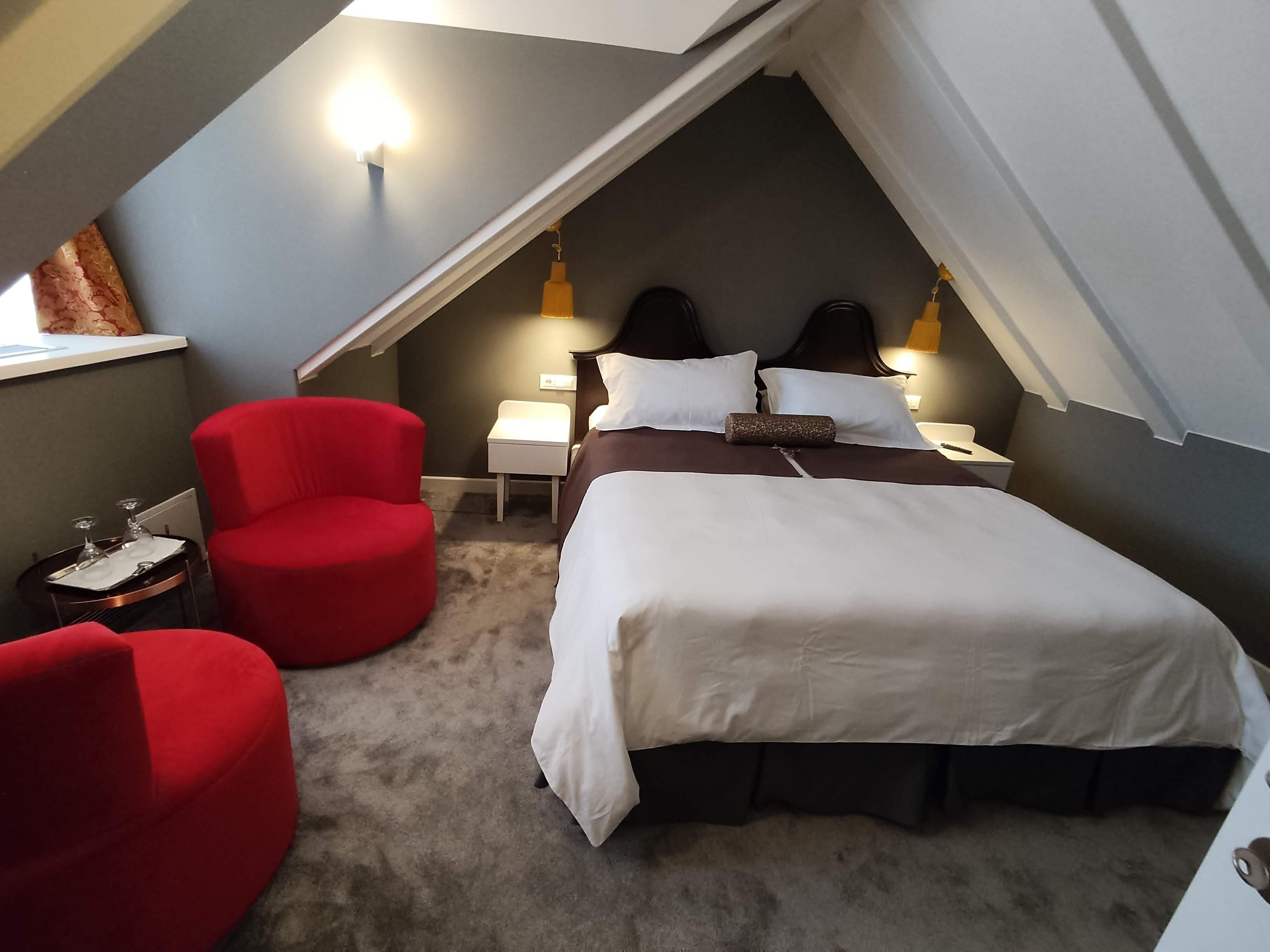 A bedroom with a bed, two nightstands, a red armchair, and a small table with glasses.