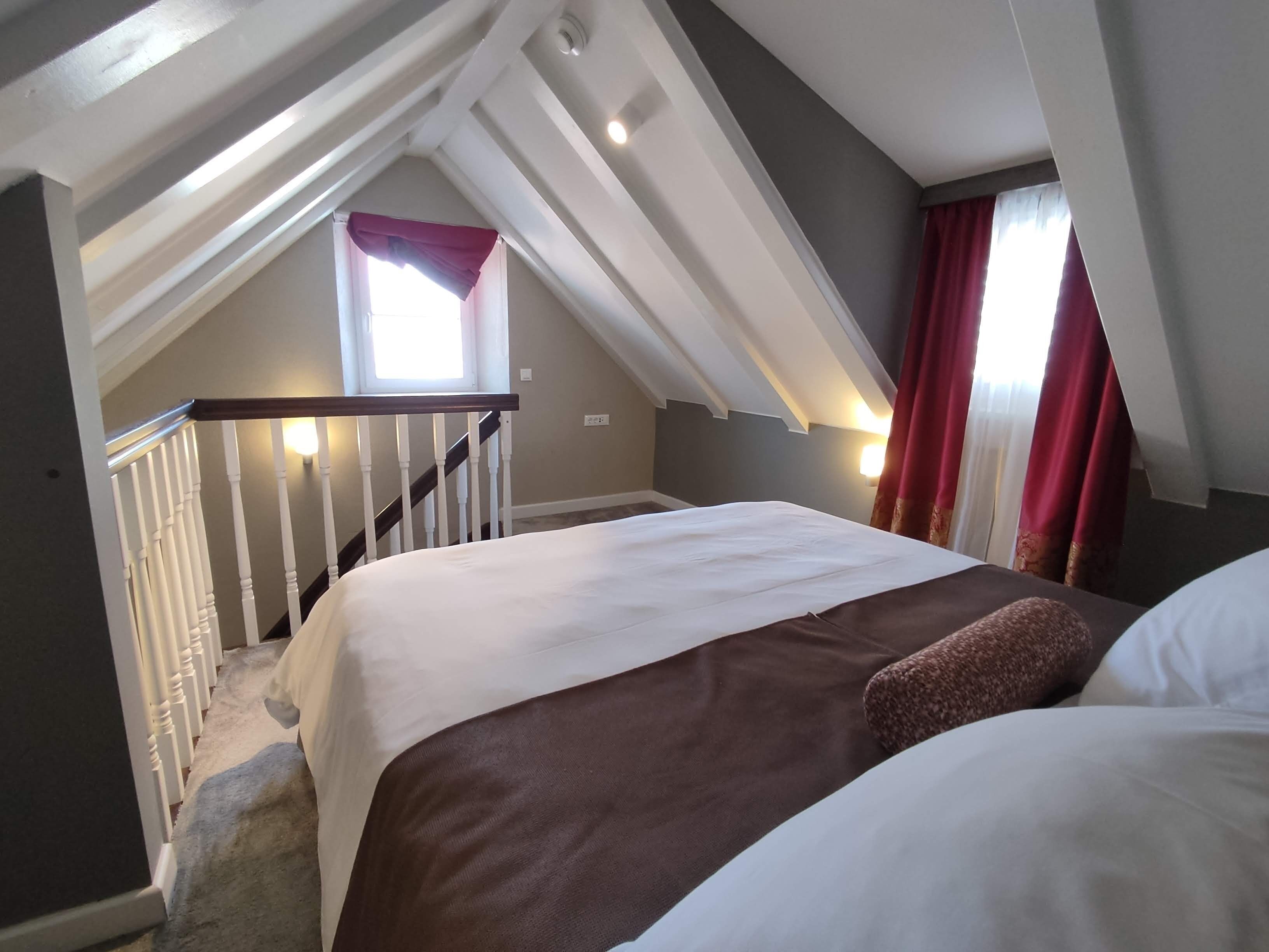 A bedroom with a sloped ceiling, a bed with white and brown bedding, a window with red curtains, and a staircase with white railings.