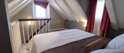 A bedroom with a sloped ceiling, a bed with white and brown bedding, a window with red curtains, and a staircase with white railings.
