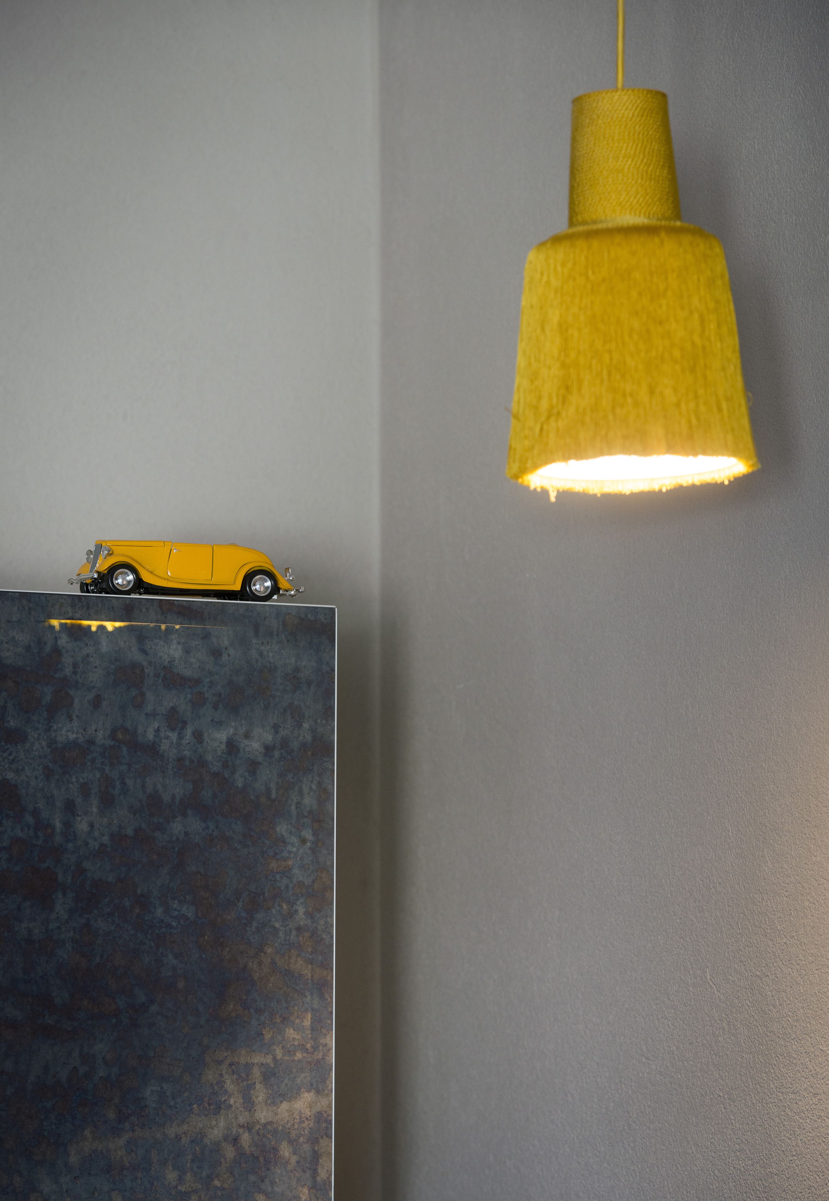 Una lámpara colgante amarilla en el techo, un pequeño coche modelo amarillo sobre una pared texturizada, y una pared lisa al fondo.