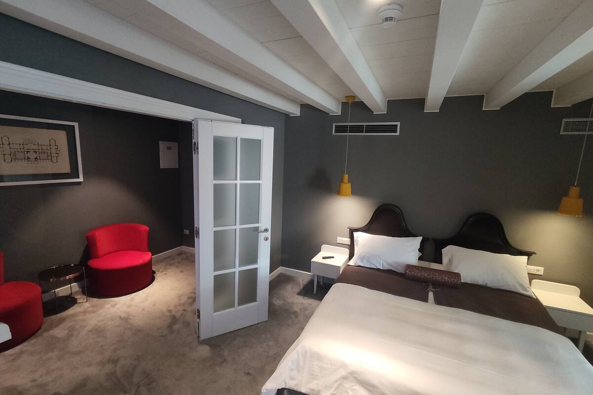 A hotel room with a bed, a red chair, a white door, and a ceiling with exposed beams.