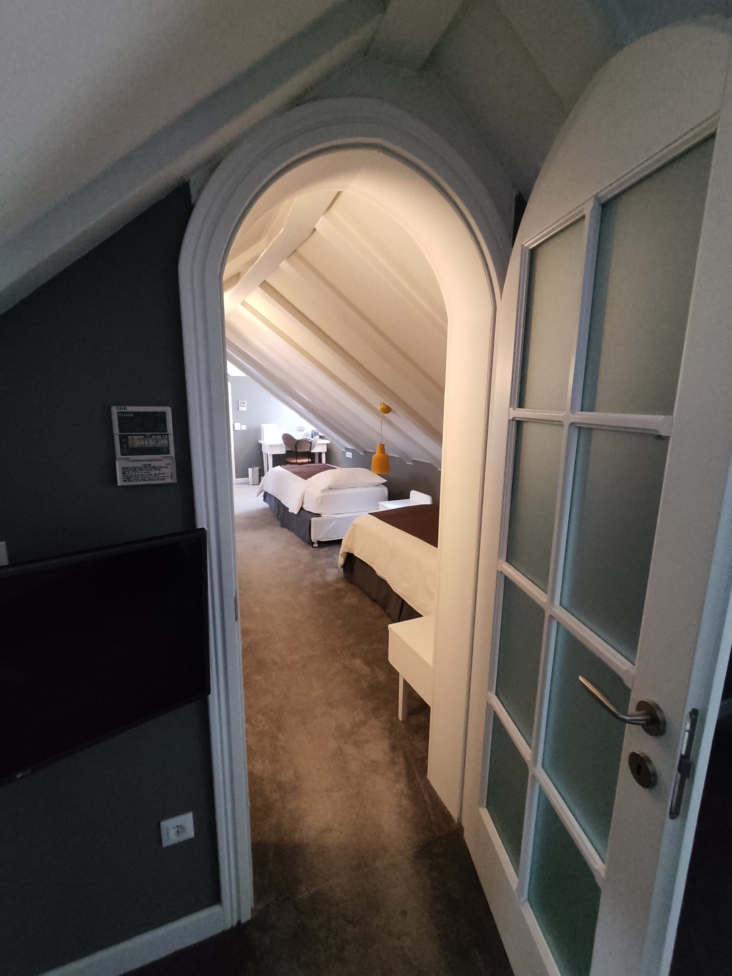 Um corredor estreito que leva a um quarto com teto inclinado, uma cama e uma escrivaninha.