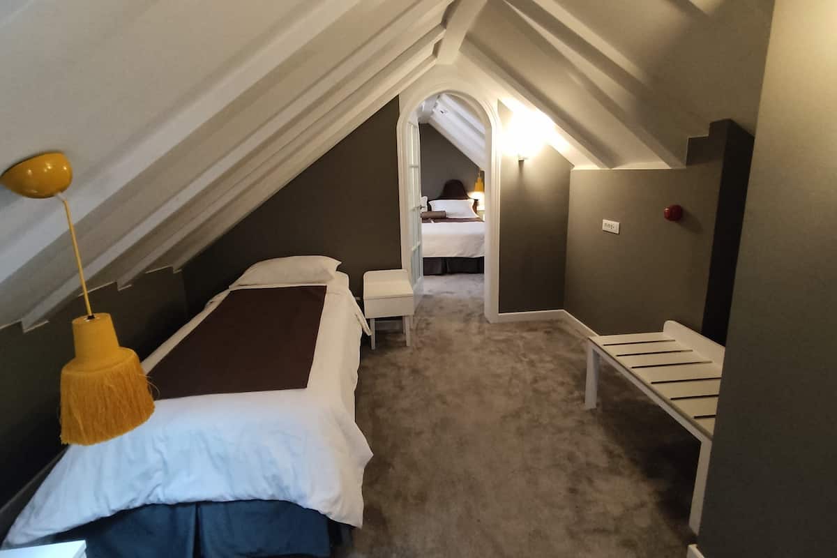 A bedroom with a sloped ceiling, a bed with white and brown bedding, a small bedside table, a yellow lamp, and a view into another room with a bed and a nightstand.