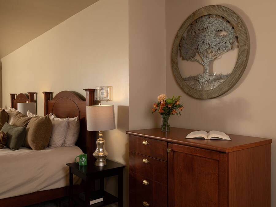 Una habitación de hotel con cama, mesita de noche, lámpara y un cuadro decorativo de un árbol.