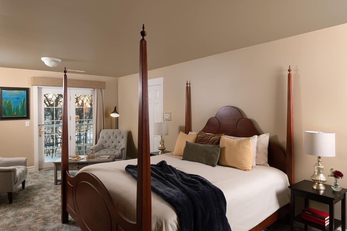 Una cama con dosel con un edredón azul oscuro, una mesita de noche de madera con una lámpara, y una silla en una habitación de hotel con vistas al exterior.