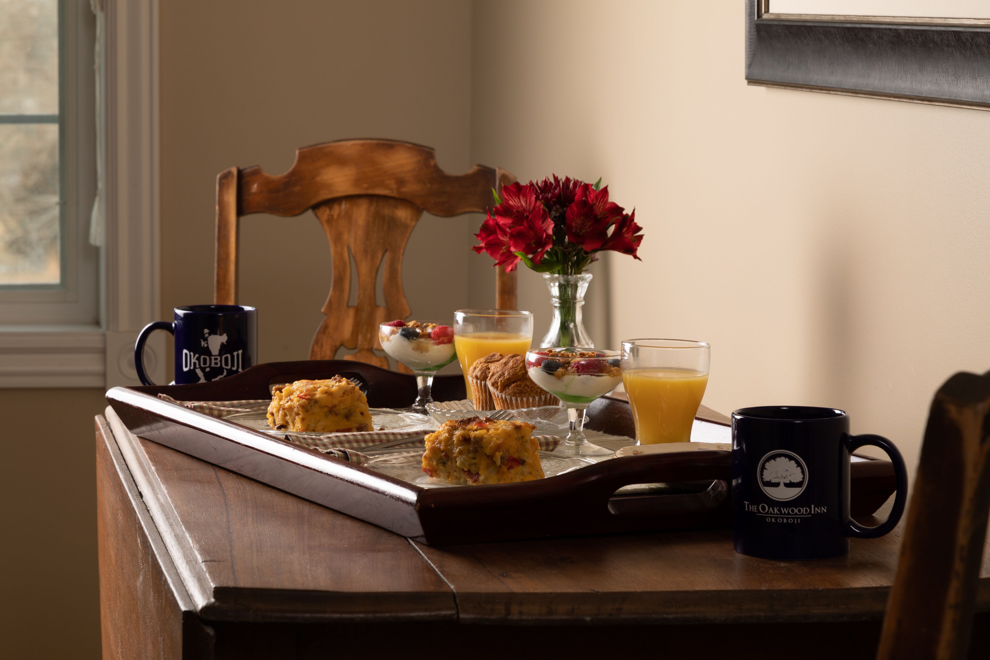 Una bandeja de desayuno con muffins, una taza, un vaso de jugo de naranja y un florero sobre una mesa de madera.