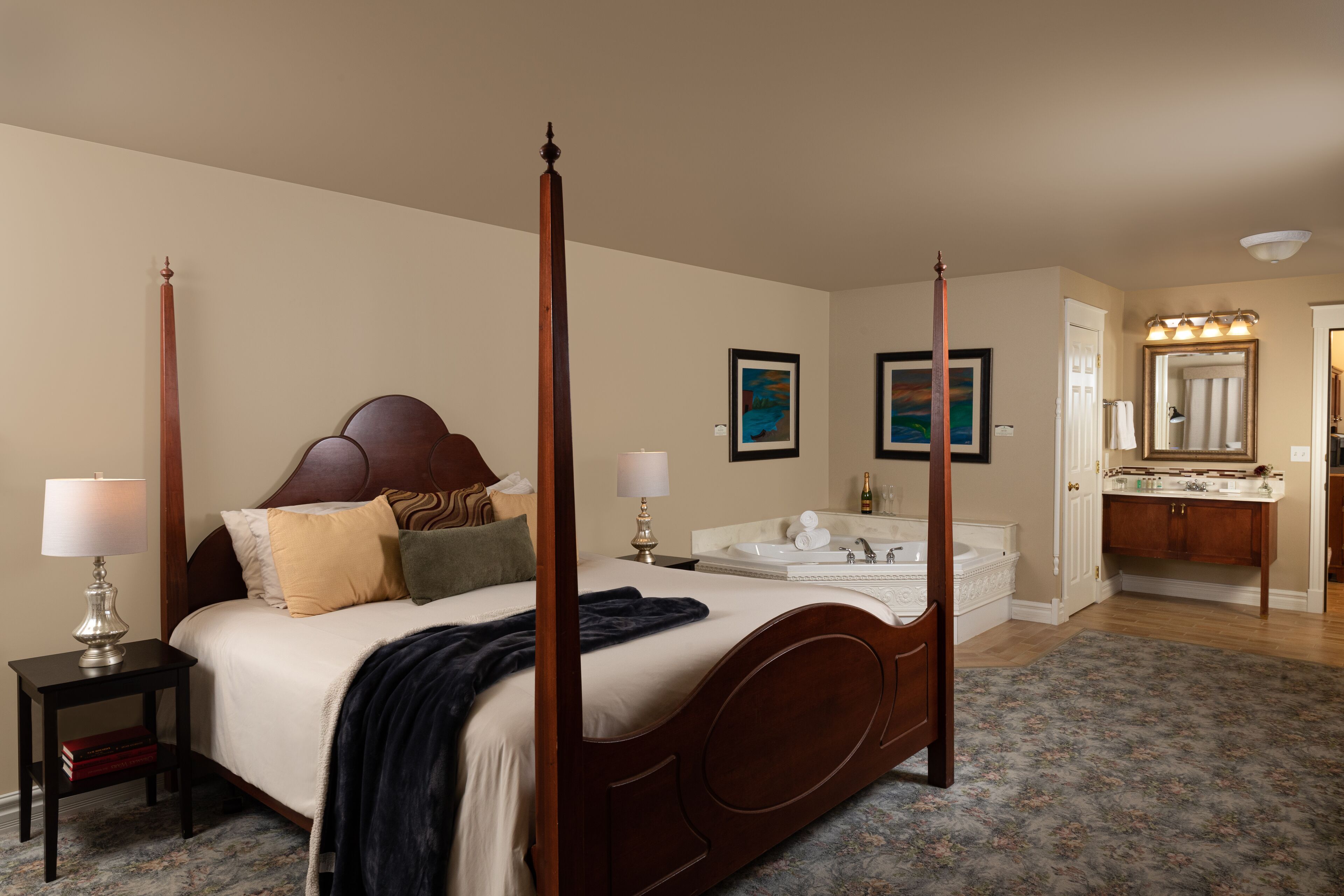 A four-poster bed with a headboard, bedside tables with lamps, a large mirror, a bathroom with a sink and vanity, and a floral-patterned rug.