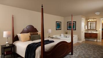 A four-poster bed with a headboard, bedside tables with lamps, a large mirror, a bathroom with a sink and vanity, and a floral-patterned rug.