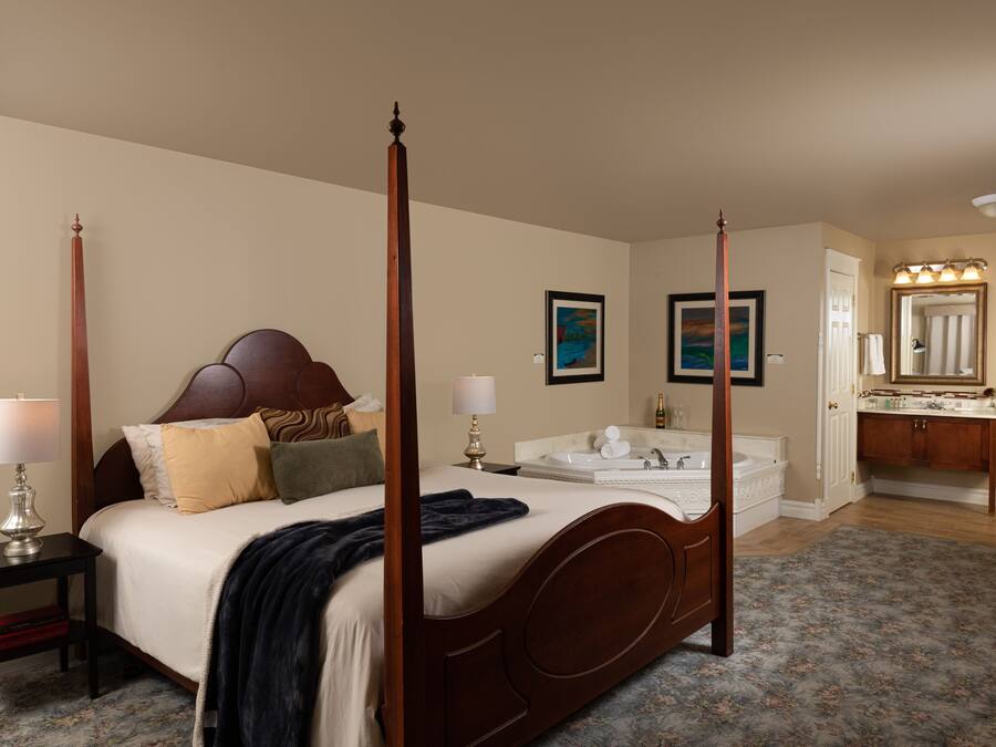 A four-poster bed with a headboard, bedside tables with lamps, a large mirror, a bathroom with a sink and vanity, and a floral-patterned rug.
