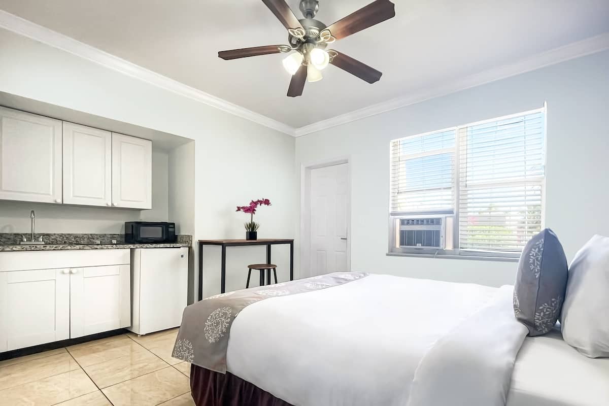 Une chambre comprenant un lit, un bureau, une chaise, un ventilateur de plafond, une fenêtre avec des stores et un coin cuisine avec des placards et un four à micro-ondes.