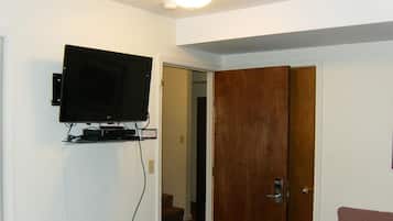 A room with a mounted television on the wall, a wooden door, and a visible staircase leading to another room.