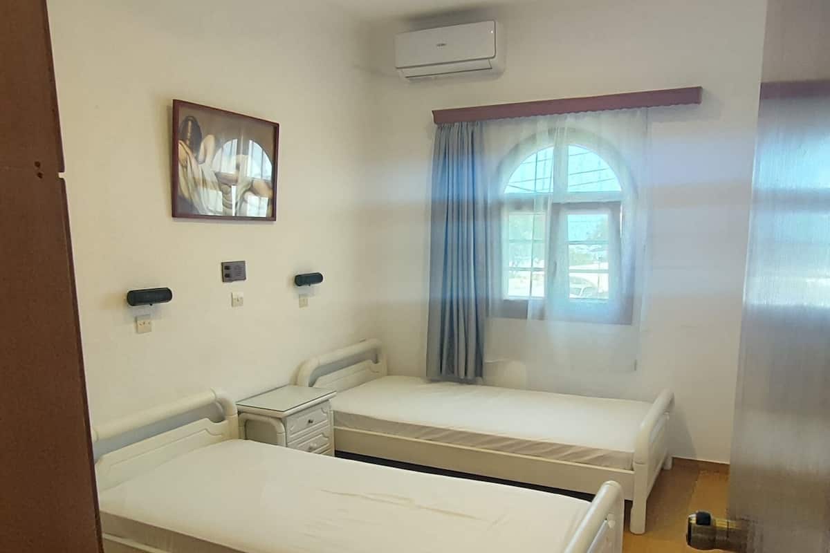 A room with two single beds, a chair, a painting, an air conditioning unit, and a window with curtains.